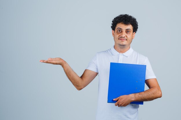 Hombre joven en camiseta blanca y jeans sosteniendo la carpeta de archivos, estirando la palma hacia afuera y mirando feliz, vista frontal.