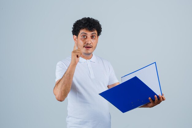 Hombre joven con camiseta blanca y jeans sosteniendo la carpeta de archivos, levantando el dedo índice en gesto eureka y mirando sensible, vista frontal.