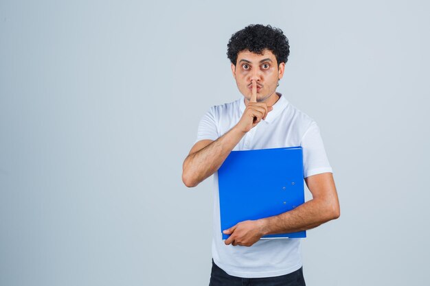 Hombre joven en camiseta blanca y jeans sosteniendo la carpeta de archivos y mostrando gesto de silencio y mirando serio, vista frontal.