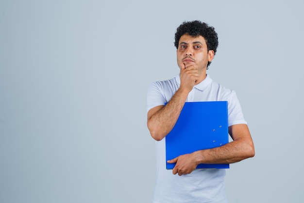 Hombre joven en camiseta blanca y jeans sosteniendo la carpeta de archivos, de pie en pose de pensamiento y mirando pensativo, vista frontal.