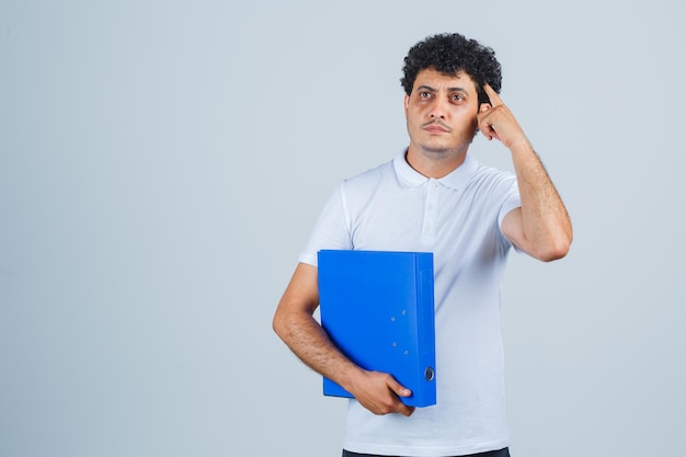 Hombre joven en camiseta blanca y jeans sosteniendo la carpeta de archivos y poniendo el dedo en la sien y mirando pensativo, vista frontal.