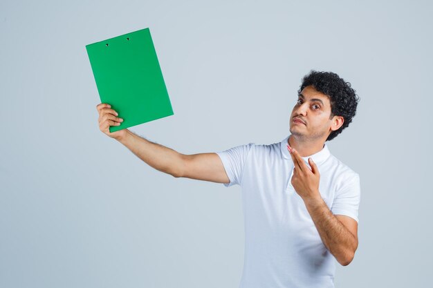 Hombre joven en camiseta blanca y jeans sosteniendo cuaderno y bolígrafo, pensando en algo y mirando pensativo, vista frontal.