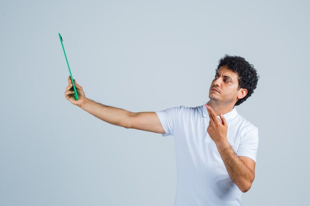 Hombre joven en camiseta blanca y jeans sosteniendo cuaderno y bolígrafo, pensando en algo y mirando pensativo, vista frontal.