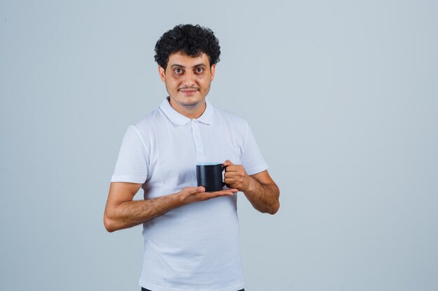 Hombre joven en camiseta blanca y jeans sosteniendo una taza de té con ambas manos y mirando feliz, vista frontal.