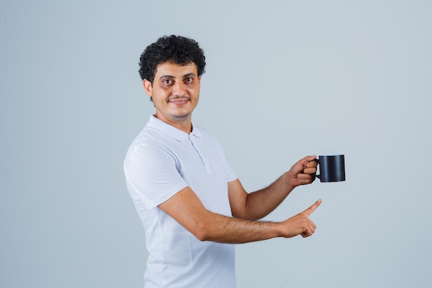 Hombre joven en camiseta blanca y jeans sosteniendo una taza de té mientras apunta hacia ella y mira feliz, vista frontal.