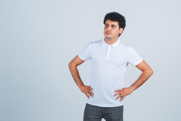 Hombre joven en camiseta blanca y jeans tomados de la mano en la cintura y mirando pensativo, vista frontal.