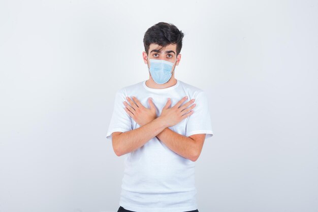 Hombre joven en camiseta blanca, máscara sosteniendo las manos cruzadas sobre el pecho y mirando confiado