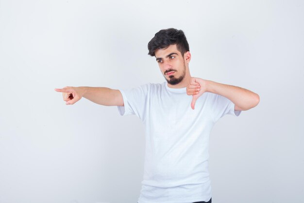 Hombre joven con camiseta blanca mostrando el pulgar hacia abajo, apuntando hacia otro lado y mirando confiado