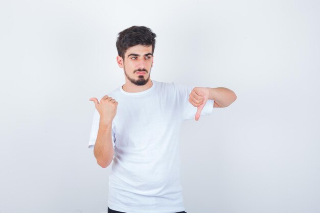 Hombre joven con camiseta blanca mostrando el pulgar hacia abajo, apuntando hacia otro lado y mirando confiado