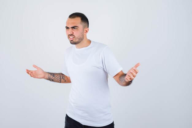 Hombre joven con camiseta blanca y pantalón negro estirando las manos de manera interrogante y mirando perplejo