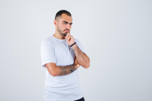 Hombre joven en camiseta blanca y pantalón negro de pie en pose de pensamiento y mirando pensativo