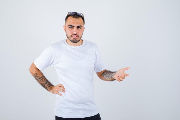 Hombre joven en camiseta blanca y pantalón negro poniendo la mano en la cintura estirando las manos como sosteniendo algo y mirando serio