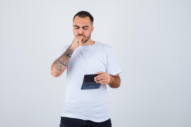 Hombre joven con camiseta blanca y pantalón negro sosteniendo la calculadora y sosteniendo el puño en la boca y mirando serio