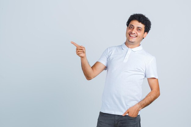 Hombre joven con camiseta blanca, pantalones apuntando a un lado y mirando feliz, vista frontal.