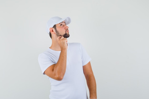 Hombre joven en camiseta blanca rascándose la barbilla y mirando pensativo, vista frontal.