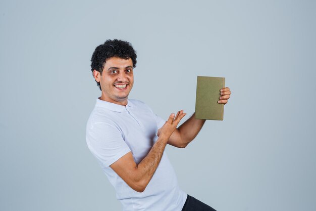 Hombre joven en camiseta blanca sosteniendo un libro y mirando alegre, vista frontal.