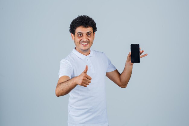 Hombre joven en camiseta blanca sosteniendo teléfono móvil, mostrando el pulgar hacia arriba y mirando alegre, vista frontal.