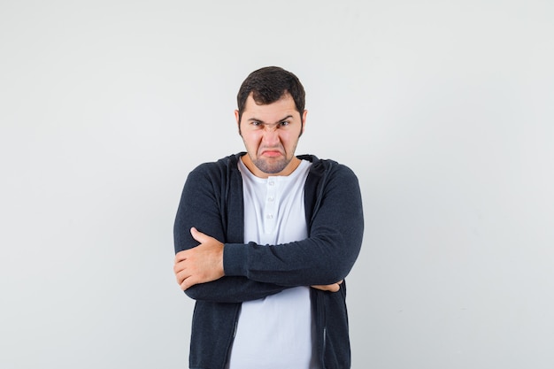 Foto gratuita hombre joven con camiseta blanca y sudadera con capucha negra con cremallera frontal de pie con los brazos cruzados y mirando enojado, vista frontal.