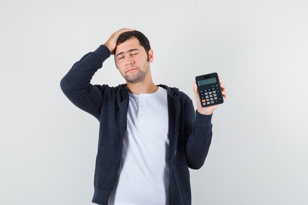 Hombre joven con camiseta blanca y sudadera con capucha negra con cremallera frontal que sostiene la calculadora y pone la mano en la cabeza y se ve optimista, vista frontal.