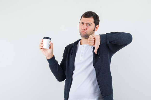 Hombre joven con camiseta blanca y sudadera con capucha negra con cremallera frontal sosteniendo una taza de café para llevar y mostrando el pulgar hacia abajo y mirando disgustado, vista frontal.