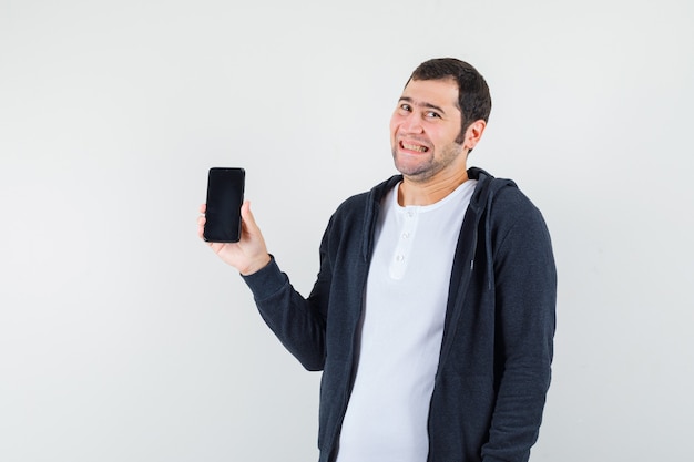 Hombre joven en camiseta blanca y sudadera con capucha negra con cremallera frontal sosteniendo teléfono inteligente, sonriendo y mirando feliz, vista frontal.