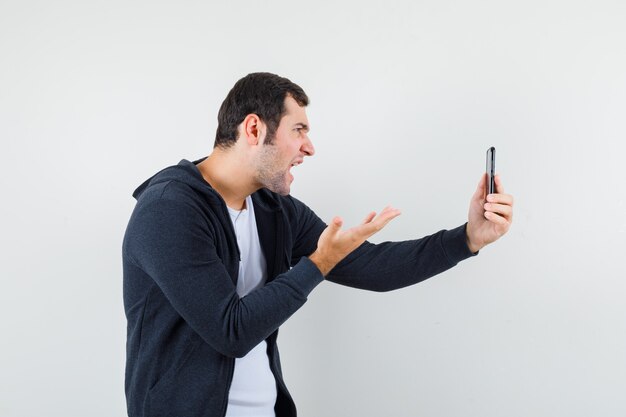 Hombre joven con camiseta blanca y sudadera con capucha negra con cremallera en la parte delantera hablando con alguien a través de una videollamada y mirando enojado, vista frontal.