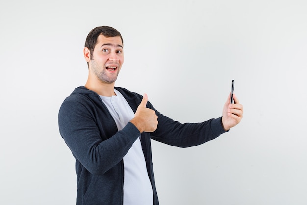 Foto gratuita hombre joven con camiseta blanca y sudadera con capucha negra con cremallera en la parte delantera que sostiene el teléfono inteligente, mostrando el pulgar hacia arriba y mirando feliz, vista frontal.