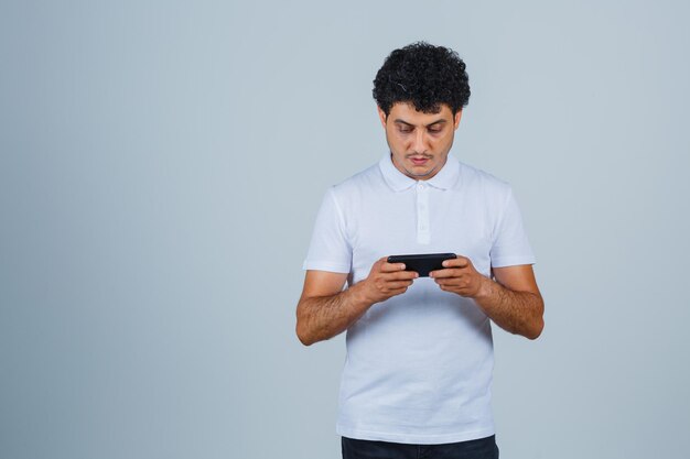 Hombre joven en camiseta blanca con teléfono móvil y mirando enfocado, vista frontal.