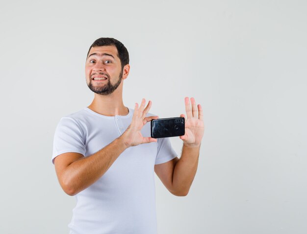 Hombre joven en camiseta blanca tomando fotos con teléfono móvil y mirando contento, vista frontal.