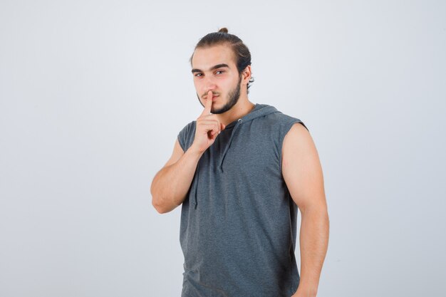 Hombre joven en camiseta con capucha mostrando gesto de silencio y mirando serio, vista frontal.