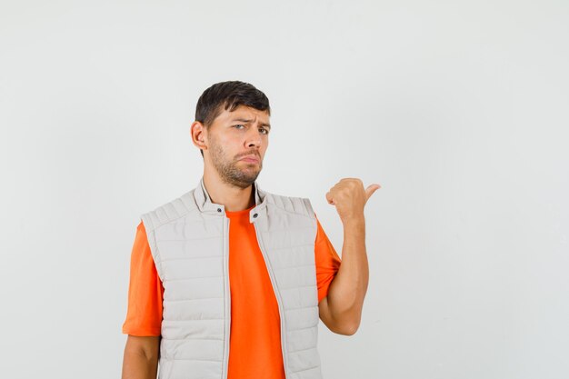 Hombre joven en camiseta, chaqueta apuntando con el pulgar hacia afuera y mirando ofendido, vista frontal.