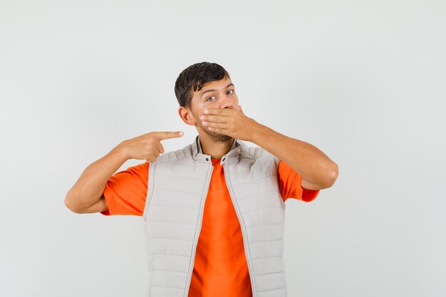 Hombre joven en camiseta, chaqueta apuntando a su mano en la boca, vista frontal.