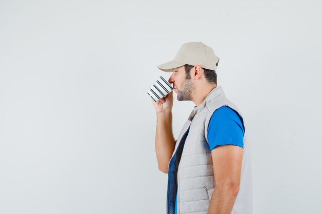 Hombre joven en camiseta, chaqueta bebiendo té aromático y mirando pensativo.