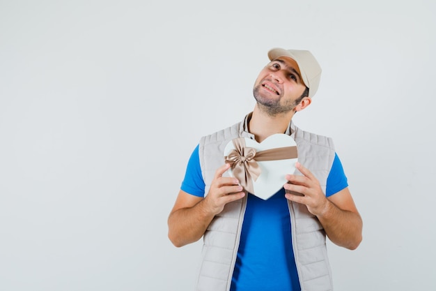 Hombre joven en camiseta, chaqueta con caja de regalo y mirada soñadora, vista frontal.