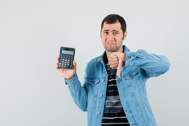 Hombre joven en camiseta, chaqueta con calculadora, mostrando el pulgar hacia abajo y mirando decepcionado