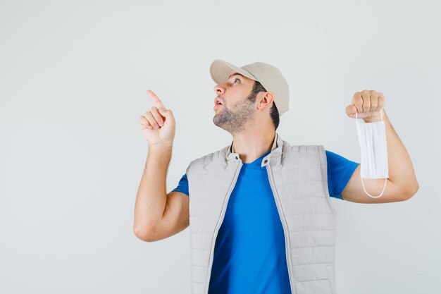 Hombre joven en camiseta, chaqueta, gorra apuntando hacia arriba, sosteniendo una máscara médica y mirando sorprendido, vista frontal.