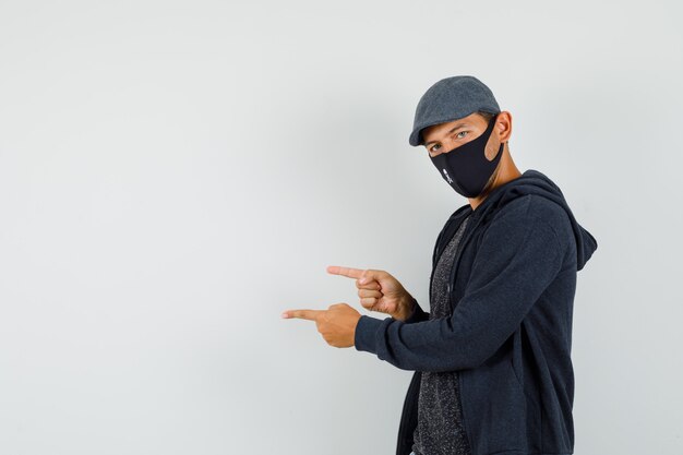 Hombre joven en camiseta, chaqueta, gorra, máscara apuntando hacia un lado.