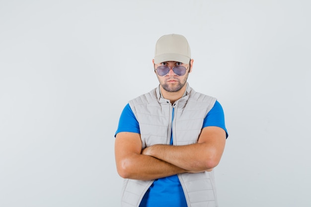 Hombre joven en camiseta, chaqueta, gorra de pie con los brazos cruzados y mirando confiado, vista frontal.