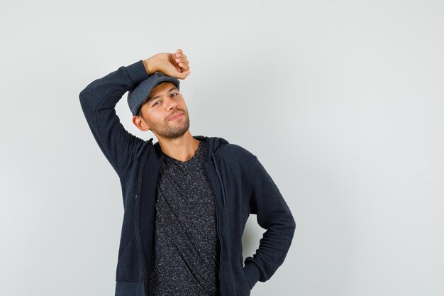 Hombre joven en camiseta, chaqueta, gorra sosteniendo el brazo levantado sobre la cabeza y luciendo guapo, vista frontal.