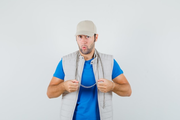 Hombre joven en camiseta, chaqueta, gorra sosteniendo su collar de cadena y mirando asombrado, vista frontal.