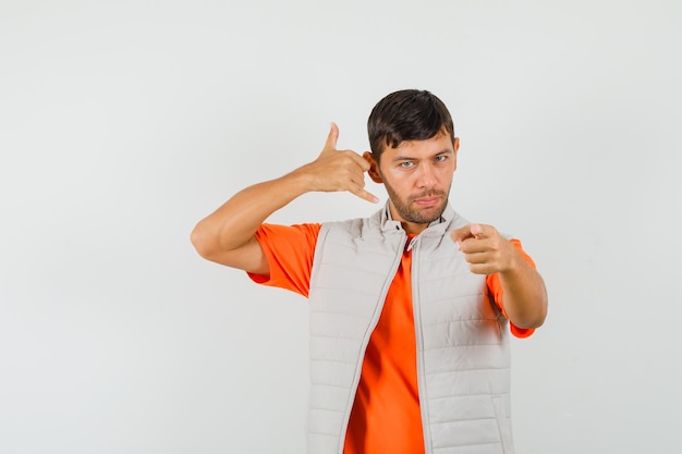 Hombre joven en camiseta, chaqueta mostrando gesto de teléfono mientras le apunta, vista frontal.