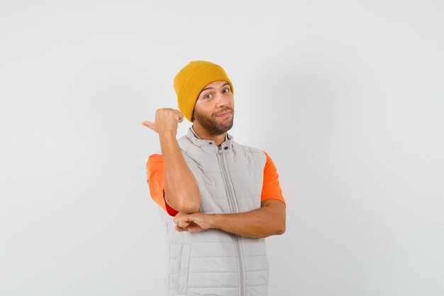 Hombre joven en camiseta, chaqueta, sombrero apuntando con el pulgar hacia atrás