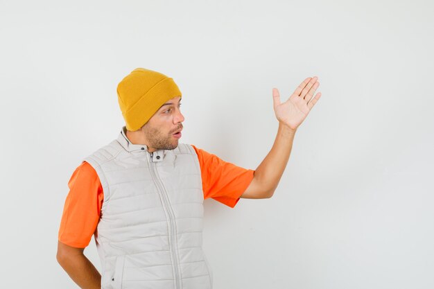 Hombre joven en camiseta, chaqueta, sombrero discutiendo algo y mirando preocupado, vista frontal.