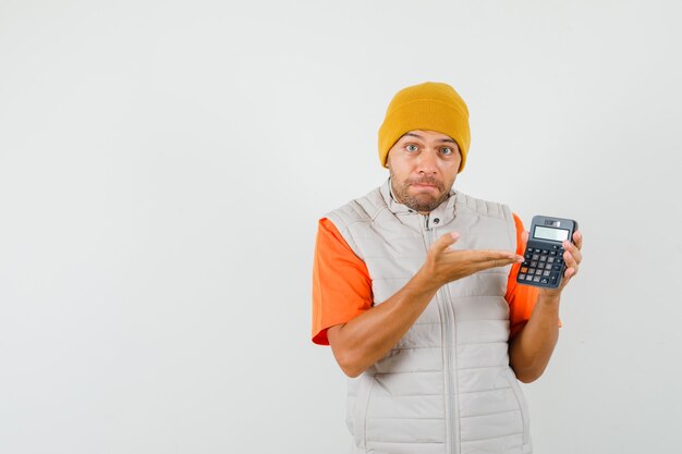 Hombre joven en camiseta, chaqueta, sombrero mostrando calculadora y mirando indefenso, vista frontal.