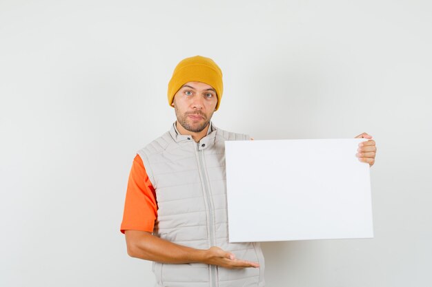 Hombre joven en camiseta, chaqueta, sombrero mostrando lienzo en blanco y mirando positivo