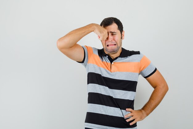 Hombre joven en camiseta frotándose los ojos mientras llora y se ve triste