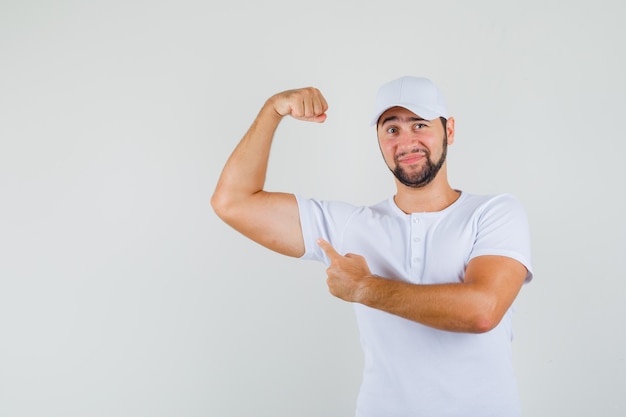 Hombre joven en camiseta, gorra apuntando al músculo de su brazo y mirando satisfecho de sí mismo, vista frontal.