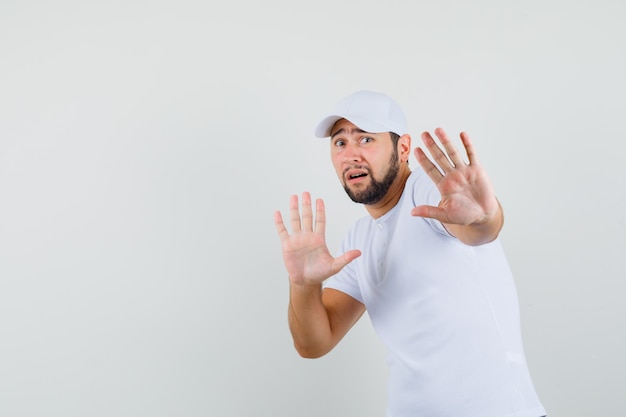 Hombre joven en camiseta, gorra levantando las manos de manera preventiva y mirando asustado, vista frontal. espacio para texto