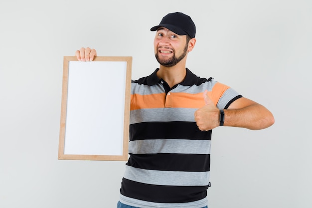 Foto gratuita hombre joven en camiseta, gorra sosteniendo un marco en blanco, mostrando el pulgar hacia arriba y mirando contento, vista frontal.