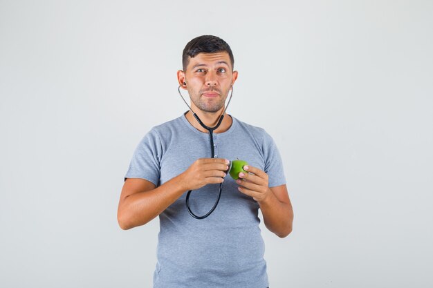 Hombre joven en camiseta gris examinando manzana verde con estetoscopio y mirando con cuidado